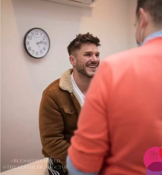 A smiling man with short hair and stubble is seated and looking to his right, engaging in conversation with Dr Ahmed after a skin care consultationwho is out of frame. He wears a brown jacket with a white shearling collar over a white shirt.