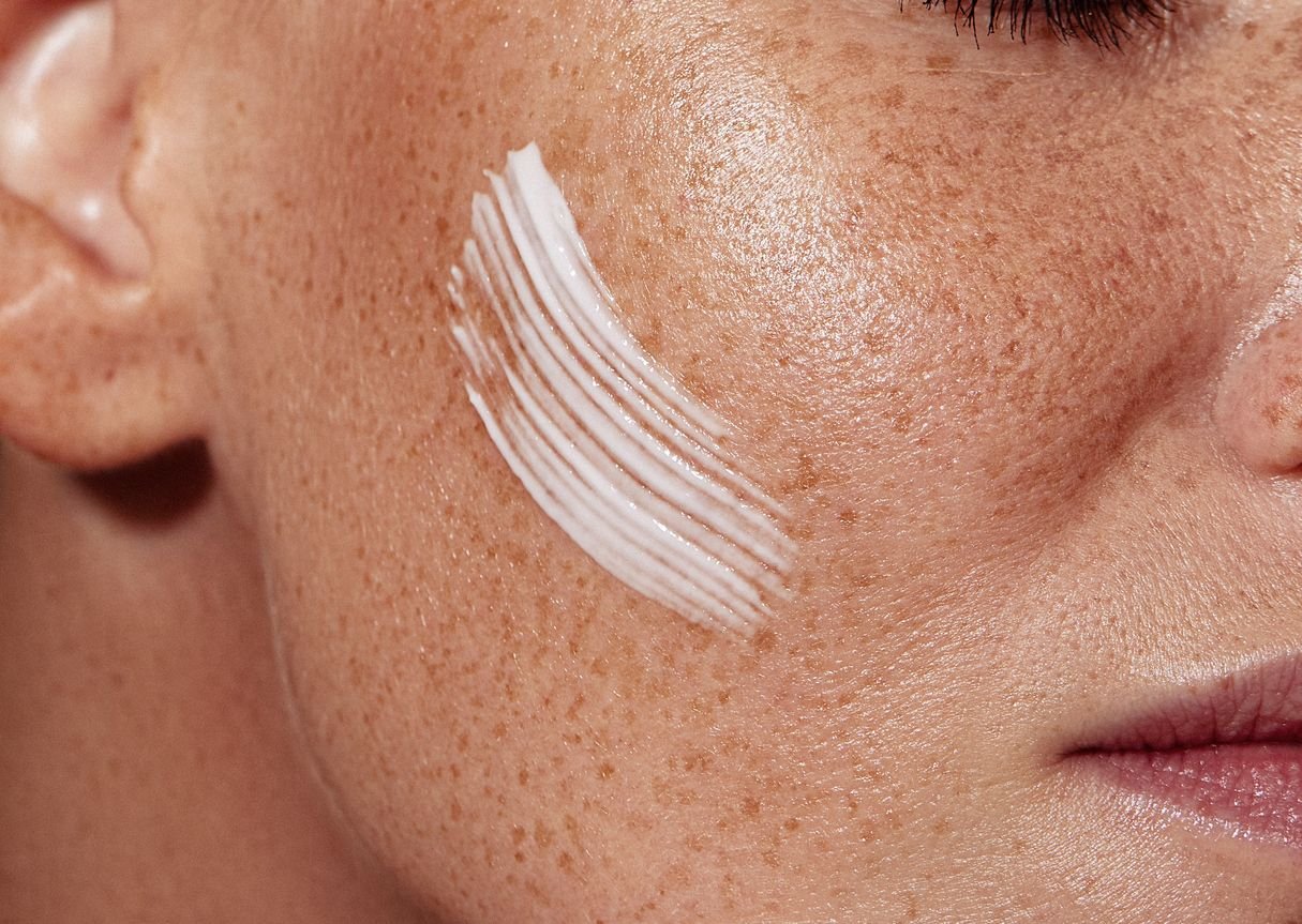 Close-up of a freckled cheek with a swipe of white skincare cream applied to the skin
